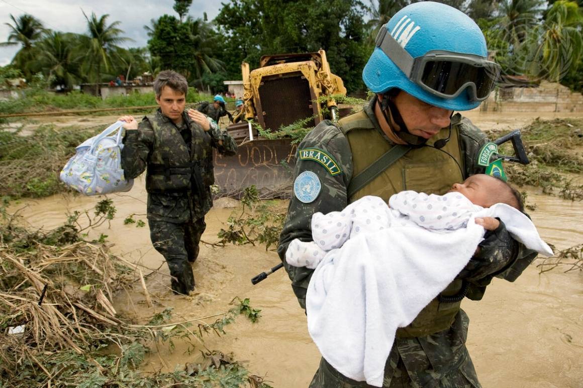 En FN-soldat bærer et spedbarn i armene, gjennom en åker oversvømmet av vann.